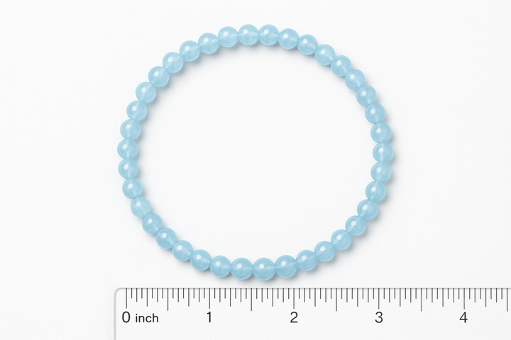 Create a realistic digital photo of a light blue aquamarine elastic beaded bracelet made of 4mm round polished beads. The bracelet has exactly 42 beads and is laid flat in a perfect circle on a white background. Include a clear inch ruler scale grid below and to the left of the bracelet for size reference. The inner diameter should be approximately 2.25 inches. The lighting should be bright and even, like a professional product image for Amazon, showing the gentle shine of the beads and accurate scale.