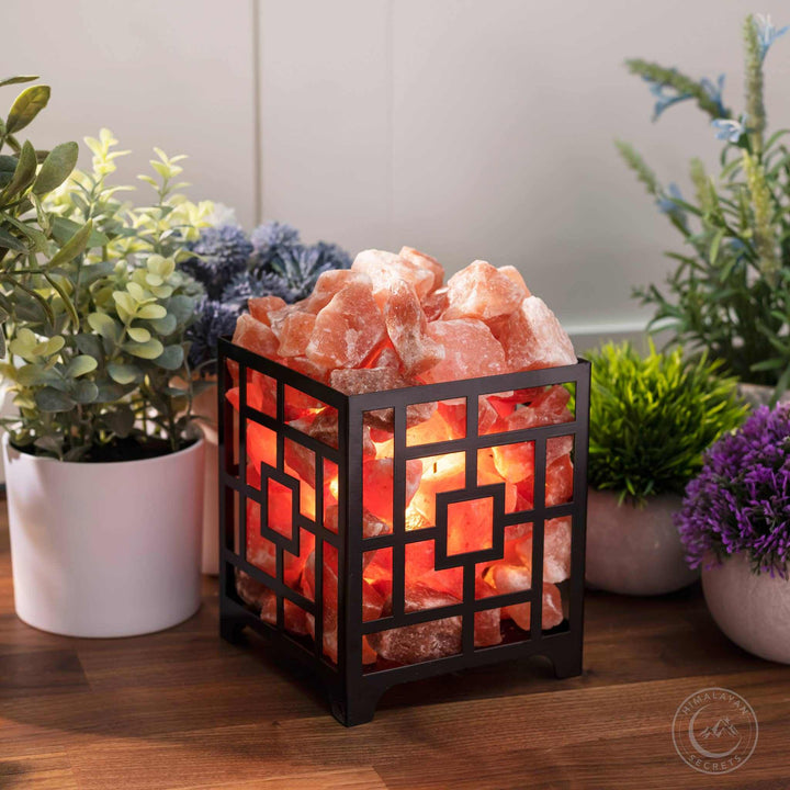 Himalayan Salt Square Basket Lamp with reddish-pink salt chunks glowing inside a black iron geometric basket on wooden surface surrounded by plants