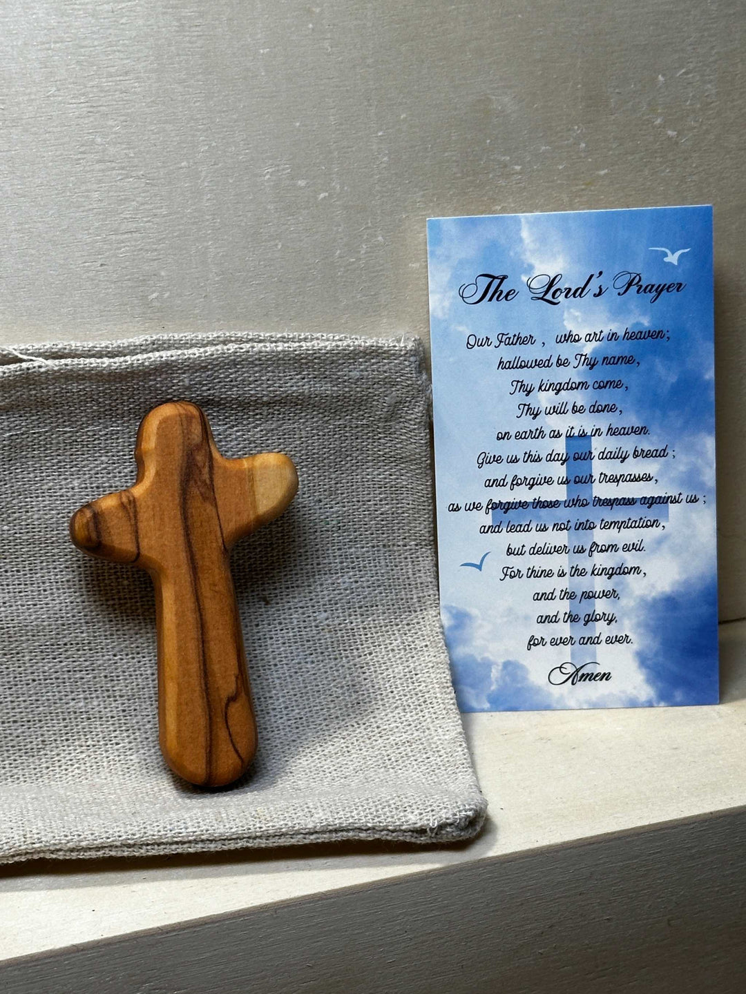 Handcrafted Olive Wood Comfort Cross with natural grain, displayed next to a printed Lord's Prayer card on a fabric surface